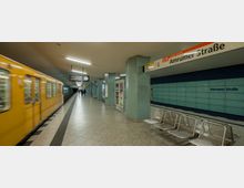U-Bahn-Station "Amrumer Straße" in Berlin mit einer gelben U-Bahn der Linie U9 auf der linken Seite, Sitzbänken aus Metall auf der rechten Seite, einer blauen Wand mit Stationsnamen im Hintergrund und türkisfarbenen Säulen in der Mitte des Bahnsteigs.