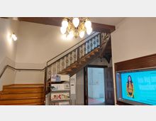 Ein Eingangsbereich mit einer Holztreppe mit verzierten Metallgeländern, einer hängenden Kronleuchterbeleuchtung und einer Wandhalterung mit Prospekten. Rechts befindet sich ein Flachbildschirm mit einer Werbeanzeige, daneben eine offene Holztür.