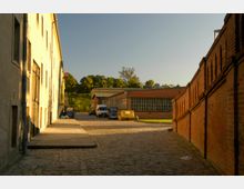 Ein gepflasterter Innenhof, umgeben von gelbem Putzgebäude links und roten Ziegelgebäuden rechts und im Hintergrund. Die Gebäude haben Fenster mit Gitterstrukturen, und im Hof stehen mehrere Fahrzeuge sowie ein gelber Container. Im Hintergrund sind Bäume vor einem klaren, blauen Himmel sichtbar.