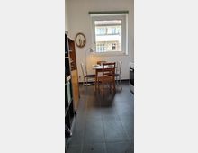 Kleine Küche mit einem runden Holztisch, umgeben von vier Stühlen, direkt unter einem Fenster mit Blick auf ein Gebäude mit Balkonen. Links steht ein Regal, rechts ein Herd, und auf der Wand hängt ein kleiner ovaler Spiegel.