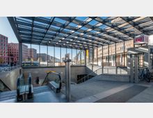 Eingangsbereich einer U-Bahn-Station mit gläsernem Dach und modernen Stahlträgern. Im Hintergrund sind mehrstöckige Bürogebäude, eine Straße mit Autos und Fahrräder sichtbar; ein DB-Logo weist auf die Deutsche Bahn hin.