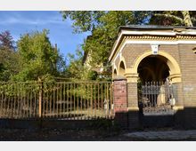 Eingang zu einem verlassenen Gebäude mit einer gelben Backsteinfassade und Rundbögen. Im Vordergrund ein rostiger Metallzaun mit Tor, dahinter dichte Vegetation.