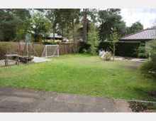 Ein Garten mit einer Rasenfläche, umgeben von Hecken und Bäumen. Im Vordergrund ist eine gepflasterte Terrasse zu sehen, rechts eine kleine Spielfläche mit einem Fußballtor und links eine Feuerstelle mit einem Holztisch.