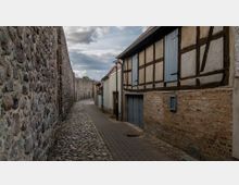 Schmale Gasse mit Kopfsteinpflaster, flankiert von einer hohen, alten Steinmauer auf der linken Seite und Fachwerkhäusern auf der rechten Seite; im Hintergrund leicht bewölkter Himmel.