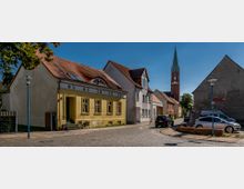 Straßenszene in einer Kleinstadt mit gepflasterter Straße, niedrigen Häusern mit Satteldächern und einem gelben Gebäude im Vordergrund. Im Hintergrund steht ein Kirchturm mit grüner Spitze, daneben geparkte Autos und ein Schild mit der Aufschrift „Museum“.