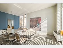 Moderner Besprechungsraum mit einem großen ovalen Tisch und mehreren beige-farbenen Bürostühlen. Der Raum hat blaue und weiße Wände, ein großes abstraktes Gemälde, bodentiefe Fenster mit Jalousien und ist von einfallendem Sonnenlicht durchzogen.