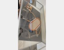 Modell eines architektonischen Entwurfs in einer Glasvitrine, das ein Gelände mit Gebäuden, Wegen und einer ovalen Anlage zeigt, umgeben von Baumreihen. Der Hintergrund ist schwarz, und erklärender Text ist auf der linken unteren Ecke zu sehen.