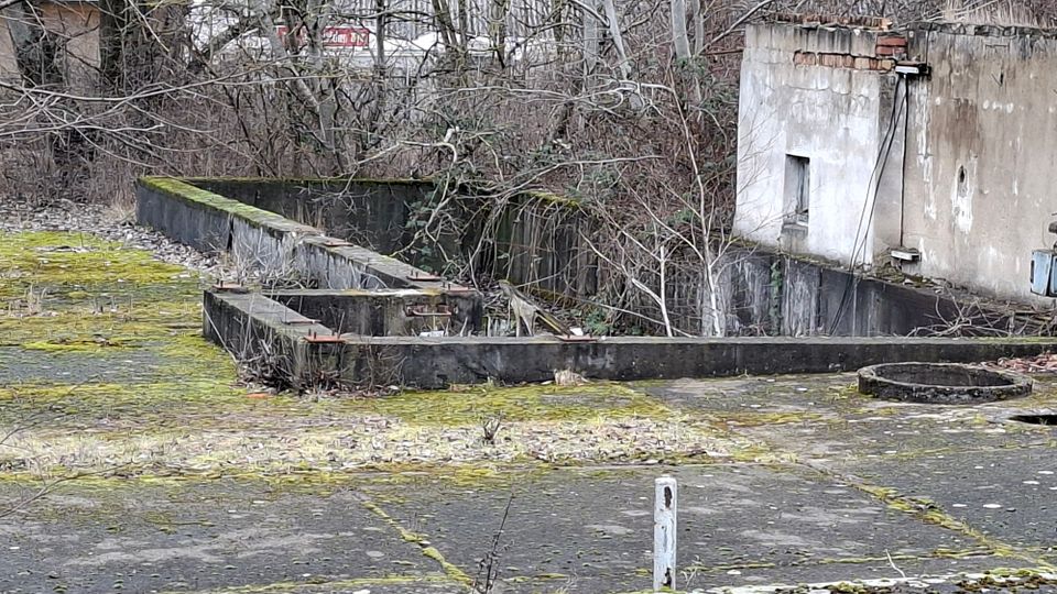 Verlassener, moosbewachsener Betonhof mit niedrigen, rechteckigen Mauern und einem runden Betonschachtdeckel. Rechts steht ein verwittertes Gebäude mit bröckelndem Putz und kleinem Fenster, im Hintergrund dichtes, überwuchertes Gehölz.