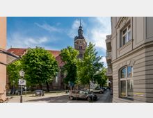 Straßenszene mit einer Kirche im Hintergrund, gekennzeichnet durch einen hohen Glockenturm mit Uhr. Im Vordergrund stehen Bäume, geparkte Autos und ein klassizistisches Gebäude mit verzierten Fassaden rechts im Bild.