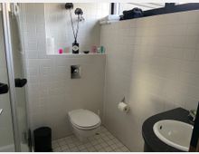 Kleines Badezimmer mit weißen Fliesen an Wänden und Boden, einer hängenden Toilette, einer schwarzen Ablage mit Waschbecken und einer Dusche mit Glastür. Über der Toilette befindet sich ein Wandregal mit Dekoration und Pflegeprodukten, daneben ein Fenster mit zusammengelegten Handtüchern.