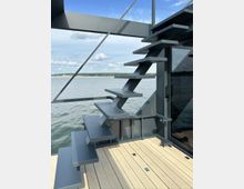 Moderne, freistehende Metalltreppe mit geradlinigem Design auf einer Terrasse aus Holzplanken, mit Blick auf einen See mit ruhiger Wasseroberfläche und bewölktem Himmel im Hintergrund.