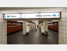 U-Bahn-Station mit weißen, gefliesten Säulen und roten Backsteinwänden; Hinweisschilder zeigen die Richtungen der Linie U7 nach „Rudow“ und „Rathaus Spandau“. Automaten stehen in der Mitte des Bahnsteigs, eine Person läuft in Richtung des Ausgangs.