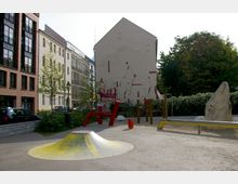 Städtischer Spielplatz mit bunten Kletterelementen und einer gelb bemalten Kuppelform im Vordergrund, umgeben von mehrstöckigen Gebäuden aus Ziegel- und Putzfassaden. Im Hintergrund ist eine bemalte Hauswand sowie grüne Büsche und Bäume zu sehen.