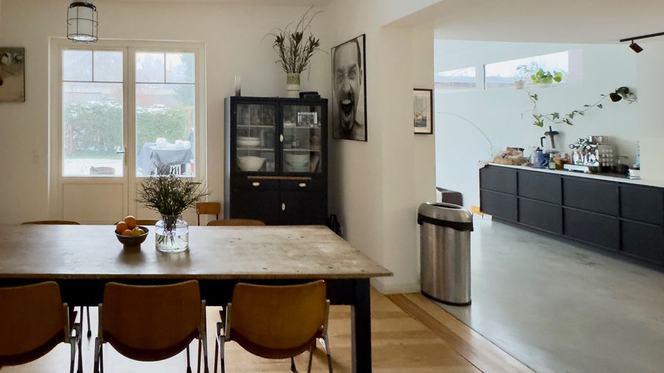 Offener Wohn- und Essbereich mit großem Holztisch und Stühlen vor einer Glastür zum Garten, daneben ein dunkler Vitrinenschrank. Rechts schließt sich eine moderne Küche mit langen, schwarzen Unterschränken, hoher Fensterzeile und Sichtbetonboden an.
