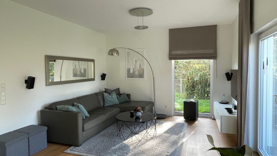 Modernes Wohnzimmer mit einer grauen Eck-Couch und mehreren blauen Kissen, einem runden schwarzen Couchtisch auf einem Teppich und einer Stehlampe mit gebogenem Arm. An einer Wand hängen ein großer Spiegel und ein Bild, gegenüber befindet sich ein Fernseher auf einem weißen Regal. Bodentiefe Fenster bieten Blick auf einen Garten.