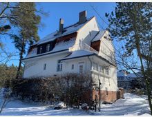 Schneeumgebenes Einfamilienhaus im Landhausstil mit weißer Fassade und rotbraunem Dach. Das Gebäude hat einen Balkon, zahlreiche Fenster und liegt in einer bewaldeten Umgebung mit kahlen Bäumen und Sträuchern unter klarem Himmel.