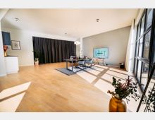 Moderner Besprechungsraum mit hellem Holzboden, großen bodentiefen Fenstern, einem schwarzen Vorhang an der Rückwand und einem Tisch mit Stühlen auf einem Teppich. Ein Monitor mit dem Schriftzug „LÖFT“ steht auf einem Stativ an der Wand.