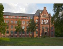 Backsteingebäude im neugotischen Stil mit großen, bogenförmigen Fenstern und spitzem Giebel, umgeben von einer Grünfläche und einzelnen Bäumen. Der Eingang hat ein verziertes Portal mit kleinen Türmen und dekorativen Elementen.