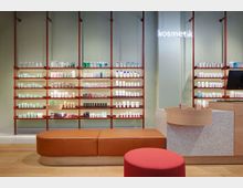 Ein moderner Verkaufsraum mit Regalen, die kosmetische Produkte in bunten Verpackungen präsentieren. Im Vordergrund befinden sich eine lederbezogene orangefarbene Sitzbank und ein roter Hocker, während rechts ein Verkaufstresen aus Holz und Steinoptik sichtbar ist.