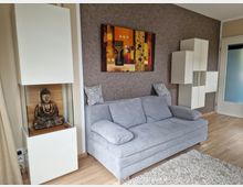 Wohnzimmer mit beige-brauner Wand, einer grauen Couch, einem Teppich aus hellem Material und modernen weißen Schränken. Links steht eine Vitrine mit einer Buddha-Statue, oben hängt ein buntes Wandbild, und rechts ist eine Tür mit mattiertem Glas zu sehen.