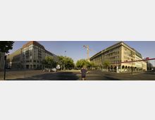 Straßenszene an einer Kreuzung in einer Stadt mit mehrstöckigen Gebäuden aus Stein und großen Fenstern. Links ein Eckgebäude mit rotem Dach, rechts ein Gebäude mit Rundbögen im Erdgeschoss; im Hintergrund Bäume, ein Kran und eine Straßenlaterne.