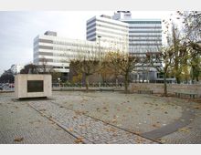 Ein gepflasterter Platz mit einer Steinplattform, die eine Gedenkflamme trägt, umgeben von Bäumen mit kahlen Ästen. Im Hintergrund stehen moderne Bürogebäude mit horizontal gestreiften Fassaden, darunter ein Gebäude mit der Aufschrift „rbb“.