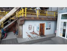 Industrielles Innenhof-Setting mit einer kleinen Sitzgruppe aus Holz und Metall vor einer grauen Wand mit Tür; eine gelbe Metalltreppe und Geländer führen nach oben, während Fahrräder und Gegenstände im Hintergrund sichtbar sind. Rechts befindet sich ein moderner Eingangsbereich mit Glastür und Aushängen.