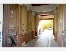 Ein überdachter Durchgang in einem alten Gebäude mit verzierten Deckenleisten und Wänden, die von Graffiti und abblätternder Farbe geprägt sind. Im Hintergrund ist ein Hof mit Bäumen und einem geparkten Auto zu sehen.