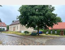 Straßenszene in einem Dorf mit Kopfsteinpflaster, einem großen Baum im Vordergrund und zwei Gebäuden im Hintergrund: ein altes Steingebäude mit verwitterter Fassade und ein rotes Ziegelhaus mit begrüntem Eingangsbereich. Ein blaues Auto steht zwischen den Gebäuden geparkt.