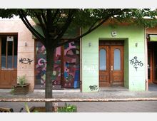 Fassade mit einer grünen und einer beige-orangefarbenen Wand, einer Holztür mit Milchglas, einem graffitibedeckten Rolltor in der Mitte und einem Baum im Vordergrund. Der Bürgersteig ist mit Kopfsteinpflaster belegt, und ein rostiges Metallgeländer trennt den Gehweg vom Straßenbereich.