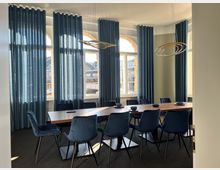 Konferenzraum mit großem Holztisch und blauen Samtstühlen, umgeben von großen Fenstern mit blauen Vorhängen. Im Hintergrund ist durch die Fenster eine städtische Szenerie zu erkennen.