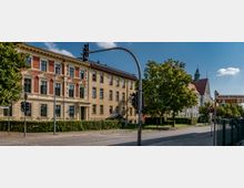 Straßenszene mit einem historischen Wohngebäude aus hellem Putz und roten Backsteinelementen links, dahinter ein großes Gebäude mit spitzem roten Dach und Turm. Im Vordergrund befinden sich eine Ampel, Straßenschilder und Bäume entlang der Straße.