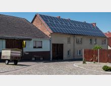 Ein ländliches Wohngebäude mit Solarzellen auf dem Dach und einer Fassade aus verputztem Mauerwerk. Im Vordergrund befinden sich ein gepflasterter Weg, ein Holzanhänger und ein Zaun aus Holzlatten.