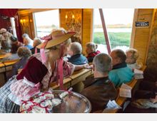 Eine Frau in historischer Kleidung mit Hut reicht einem Publikum in einem holzvertäfelten Innenraum, der vermutlich ein Boot ist, kleine verpackte Snacks auf einem Tablett. Im Hintergrund sind große Fenster mit Blick auf eine Landschaft mit Fluss und Wiesen sichtbar.