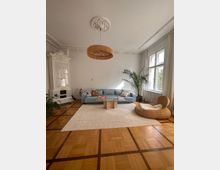 Helles Wohnzimmer mit Holzparkettboden, einer weißen Stuckdecke und einem dekorativen Kachelofen. Eine blaue Couch steht auf einem beigen Teppich, daneben stehen eine Rattansessel-Liege und mehrere große Zimmerpflanzen, während durch ein großes Fenster Tageslicht einfällt.