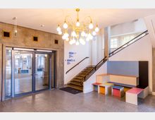 Heller Eingangsbereich mit Glastüren, steinerner Wandverkleidung und einem großen Kronleuchter mit vielen Kugelleuchten. Rechts führt eine Treppe mit Geländer nach oben; daneben stehen modulare Sitzbänke und kleine Hocker in verschiedenen Farben.