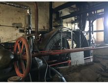 Historische Maschinenhalle mit großen Schwungrädern, Rohrleitungen und mechanischen Bauteilen in einer gealterten Industrieumgebung. Die Wände sind abgenutzt, und die Konstruktion wirkt robust und funktional.