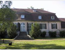 Historisches Herrenhaus mit geschwungener Fassade, kleinen Dachgauben und einem roten Ziegeldach. Vor dem Gebäude führen Stufen zu einer verglasten Eingangstür, umgeben von symmetrisch angeordneten Fenstern mit grünen Klappläden, und das Anwesen ist von Bäumen und gepflegter Rasenfläche umgeben.