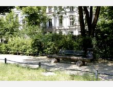 Eine Parkszene mit einer einfachen Holzbank und einem Abfalleimer rechts daneben, umgeben von grünen Büschen und Bäumen. Im Hintergrund ist ein historisches Gebäude mit weißen Fassaden und großen Fenstern zu sehen.