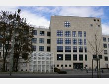 Modernes Verwaltungsgebäude mit heller Steinfassade und einem Raster aus quadratischen Fenstern. Im Vordergrund stehen kahle Bäume und eine weiße, gitterartige Skulptur aus Metall.