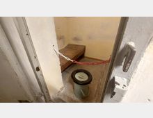 Kleiner, einfacher Gefängnisraum mit abgenutzten gelben Wänden, einer Holzpritsche und einem Eimer mit Deckel. Eine rot-weiße Kette sperrt den Zugang ab.