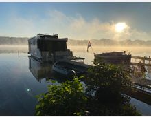 Ein Hausboot und kleine Boote liegen an einem stillen See, umgeben von leichter Nebelstimmung, während die Sonne hinter Bäumen am Horizont aufgeht. Im Vordergrund sind Pflanzen und ein kleiner Steg zu sehen, das Wasser reflektiert das Licht der Morgensonne.