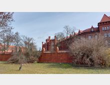 Historisches Backsteingebäude mit roten Dächern und spitzbogigen Fenstern, umgeben von einer hohen Backsteinmauer; im Vordergrund eine Grasfläche mit Büschen und kahlen Bäumen.