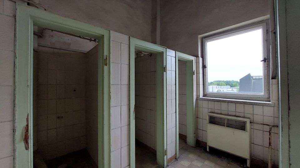 Abgenutzter Duschraum mit drei offenen Duschkabinen, weißen Wandfliesen und blassgrünen Türrahmen. Rechts befindet sich ein Fenster mit Blick auf eine Industrielandschaft sowie ein alter Heizkörper unterhalb des Fensters.