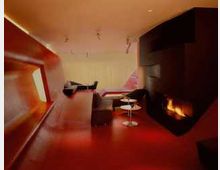 Ein modern gestalteter Innenraum mit roten Wänden und Böden, einer schwarzen Wand mit integriertem Kamin, und schlichten Sitzmöbeln. Im Hintergrund ist ein großer, heller Bereich mit einem schmalen Fenster sichtbar.