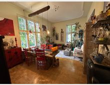 Wohnzimmer mit Essbereich in einem hellen Raum mit Holzfußboden. Im Vordergrund ein Esstisch mit roten Stühlen und Dekorationen, im Hintergrund ein Sofa, Regale, viele Pflanzen und große Fenster mit Blick ins Grüne.