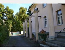 Beige Gebäude mit Säulen entlang eines Eingangsbereichs, flankiert von einer Hecke und Bäumen. Im Hintergrund steht ein grüner Müllcontainer auf einem gepflasterten Weg.