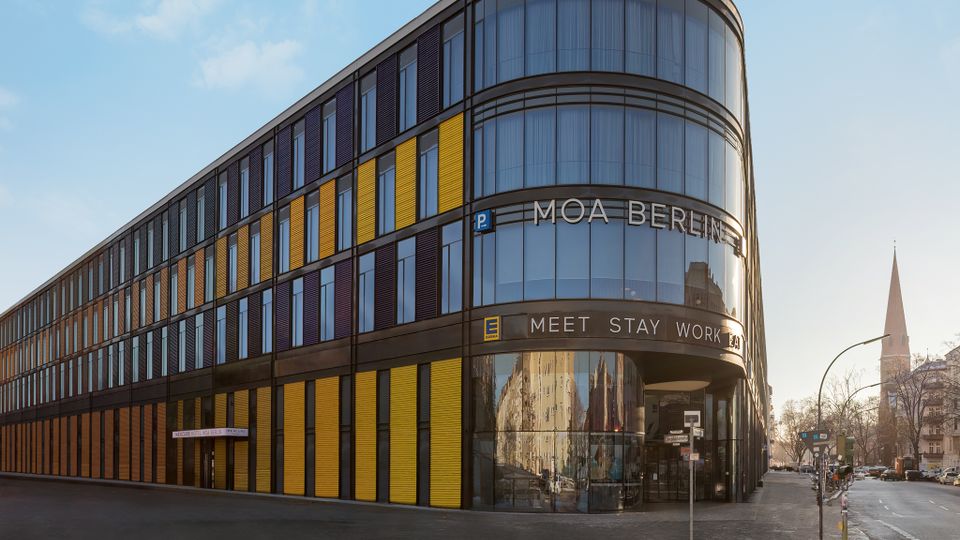 Ein modernes, mehrstöckiges Gebäude mit abgerundeter Ecke und einer Fassade aus Glas, gelben und dunklen Paneelen, beschriftet mit „MOA Berlin“. Im Hintergrund ragt ein Kirchturm in den Himmel, umgeben von Stadtgebäuden.