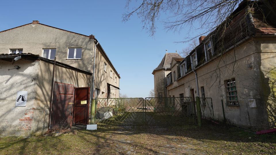 Blick in einen verwitterten Hof zwischen zwei alten, verputzten Gebäuden mit kleinen Fenstern und teils beschädigtem Dach. Ein grünes Metalltor versperrt die Einfahrt; der Boden besteht aus unregelmäßigem Kopfsteinpflaster, darüber ragen kahle Baumäste in den Himmel.
