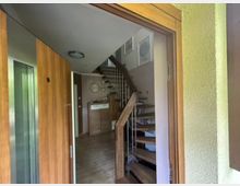 Blick durch die geöffnete Haustür in einen Wohnhausflur mit Holzrahmen, grauem Bodenbelag und einem offenen Treppenaufgang aus Holz mit Metallgeländer. Im Hintergrund stehen eine Kommode und Wanddekoration, daneben führt ein Flur zu weiteren Räumen.